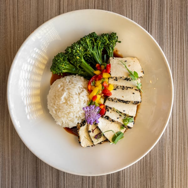 Grilled tofu with sesame seeds, rice, broccolini, mango salsa, and a purple flower on a white plate.