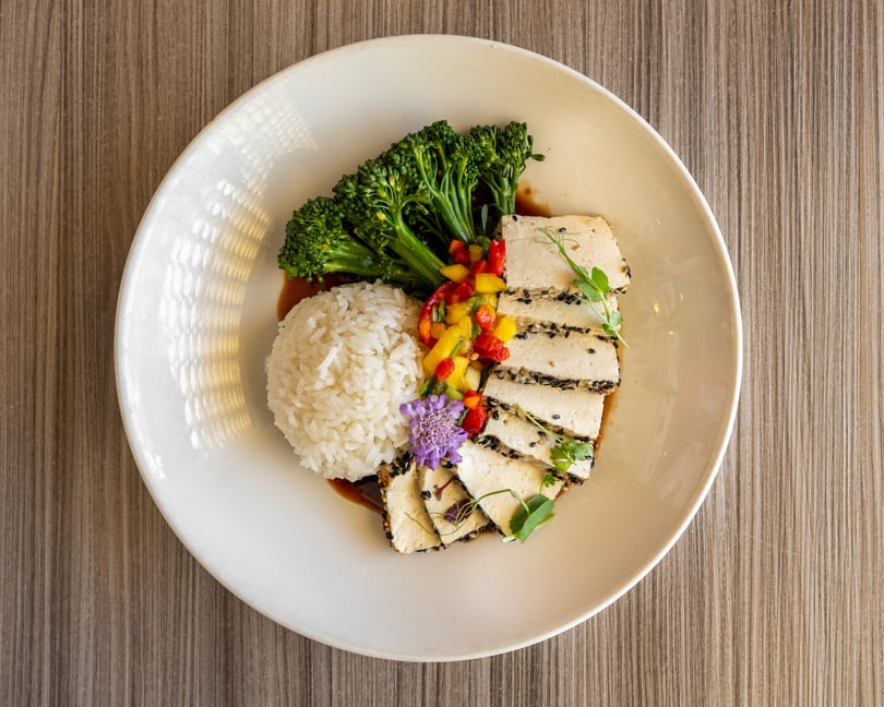 Grilled tofu with sesame seeds, rice, broccolini, mango salsa, and a purple flower on a white plate.