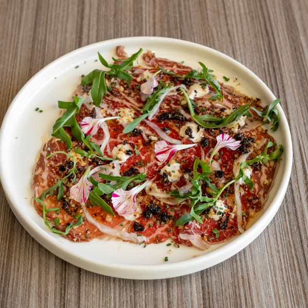 A white plate of thinly sliced meat, garnished with arugula, edible flowers, and cheese.