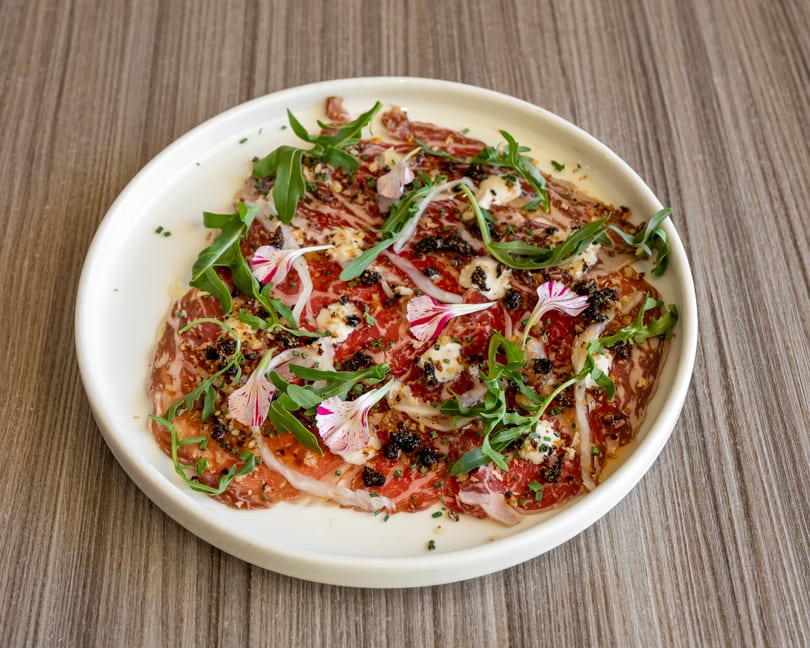 A white plate of thinly sliced meat, garnished with arugula, edible flowers, and cheese.