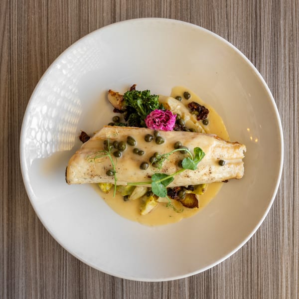 Pan-seared fish fillet with capers, broccolini, and a pink edible flower on a white plate.