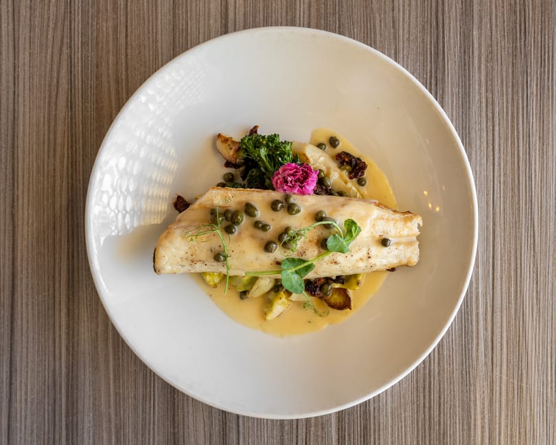 Pan-seared fish fillet with capers, broccolini, and a pink edible flower on a white plate.