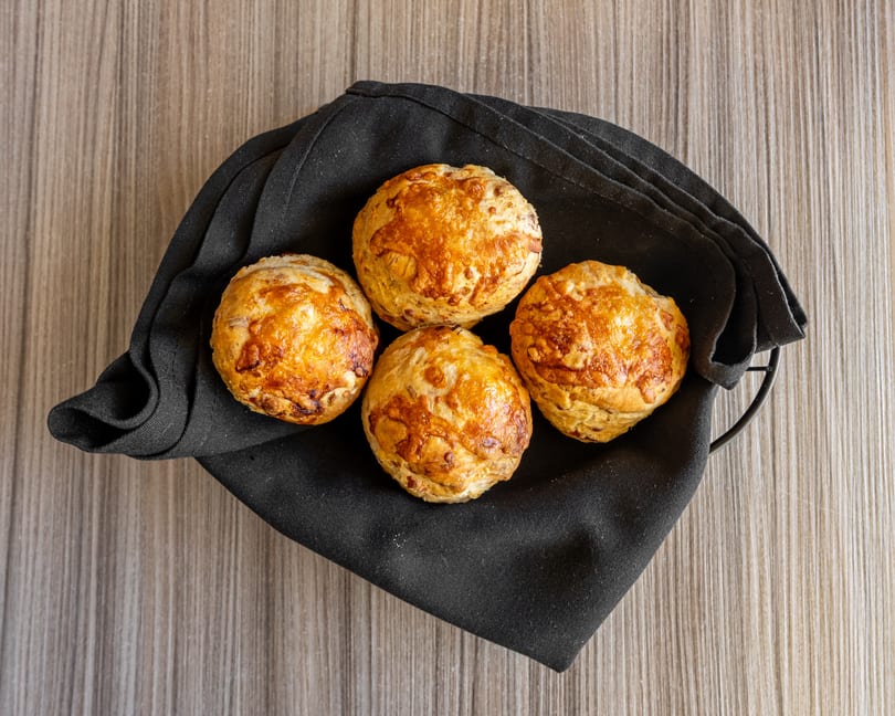 Four golden-brown cheese buns in a black napkin-lined basket on a wooden table.