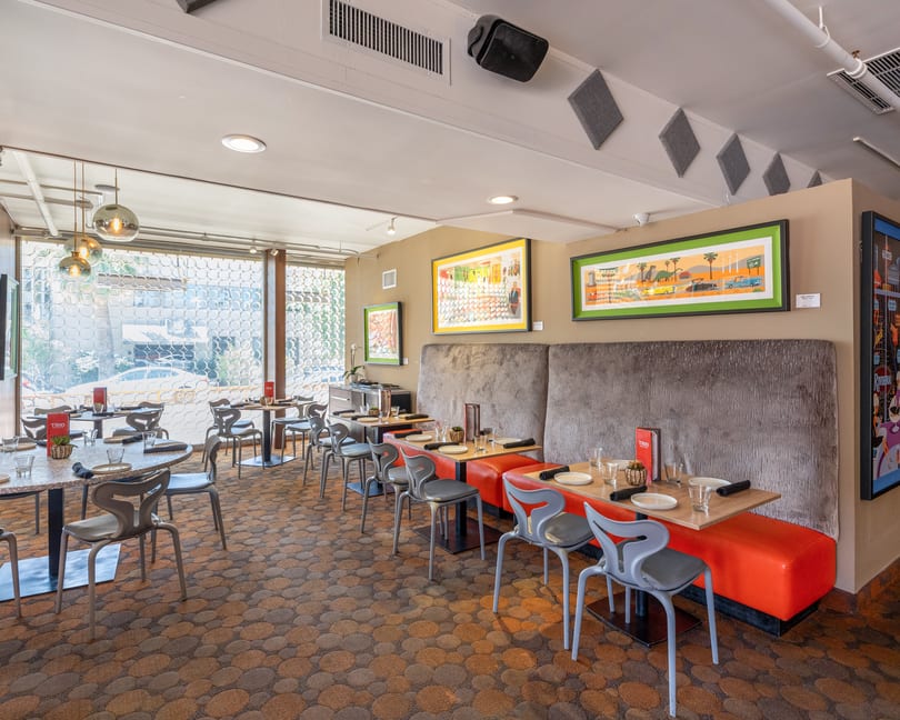 Modern restaurant interior with patterned carpet, orange booths, gray chairs, and colorful framed art on tan walls.