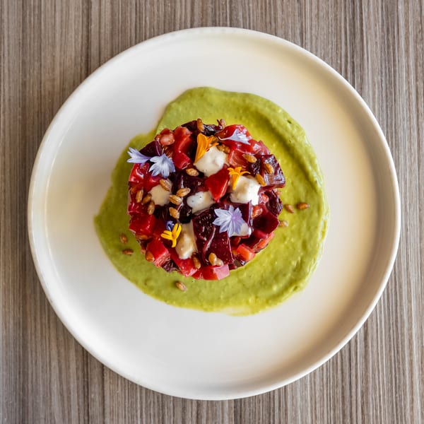 Vibrant beet tartare with creamy sauce, edible flowers, and seeds on a white plate.