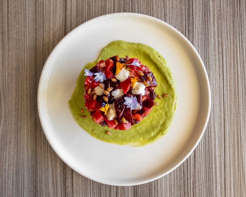 Vibrant beet tartare with creamy sauce, edible flowers, and seeds on a white plate.