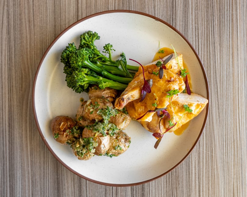 Roasted chicken with crispy skin, broccolini, and potatoes with green herb sauce on a white plate.