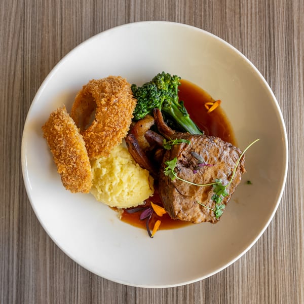 Braised beef with mashed potatoes, onion rings, broccolini, and gravy on a white plate.