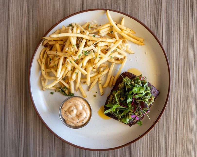 Steak frites with chimichurri, parmesan fries, and a side of creamy sauce on a white plate.